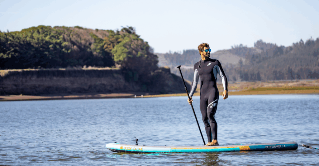 Man wearing a wetsuit and sunglasses stand-up paddling on calm cold water, surrounded by nature, with trees and hills in the background.